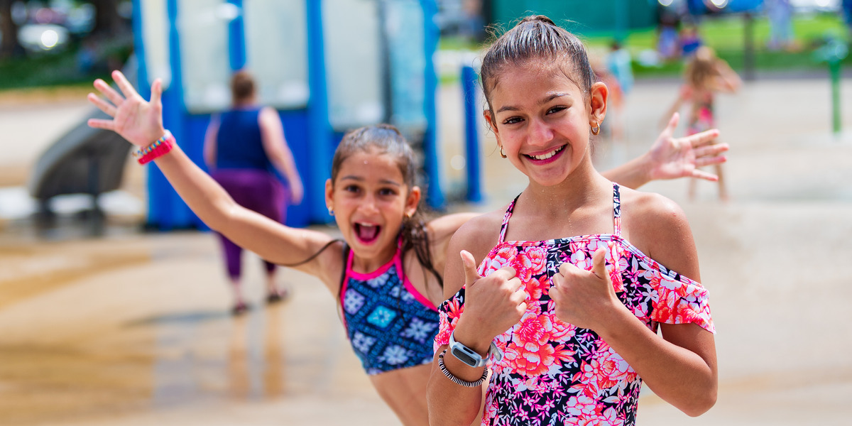 Two young girls in brightly coloured bathing suits, one posing with two thumbs up and the other behind her with her arms up in excitement in City Park Waterpark.