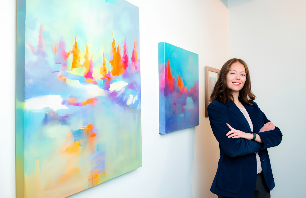 An artist wearing a navy suit stands smiling in front of a colourful painting exhibit at the Kelowna Community Theatre.