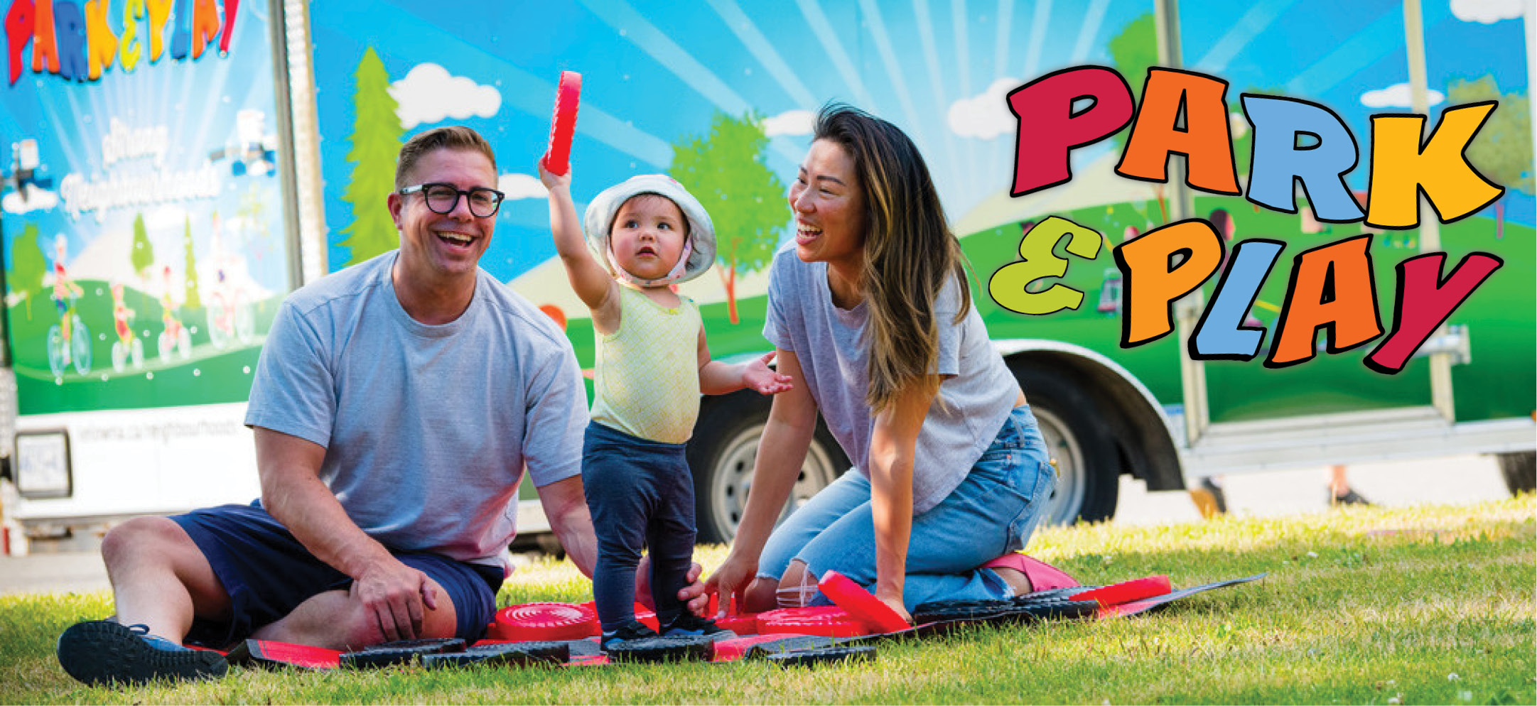 Two parents sitting on a blanket at a park on a sunny day smiling at their toddler, who is smiling, standing and holding up a giant checker piece.