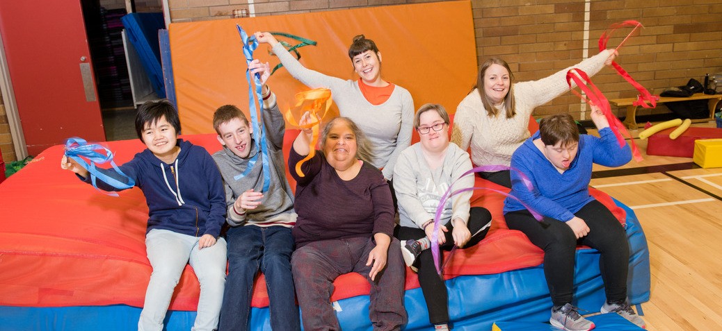 A group of friends sits on a large crash pad laughing, smiling and waving coloured ribbons