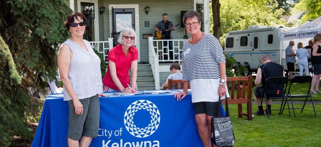 Three smiling women gather around a City of Kelowna information table