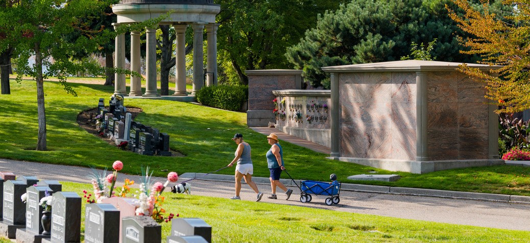 Two women walk through Kelowna Memorial Park Cemetery on a sunny day. They are pulling a blue wagon with a young child in it. The path is surrounded by memorial statues and green lawns.
