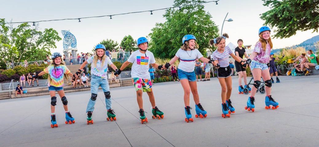 A group of young people laughing and holding hands while roller skating around at Stuart Park, wearing matching blue helmets and skates.