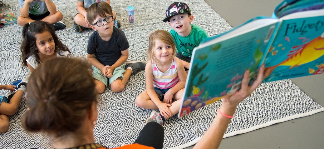 A small group of children looking up at a teacher reading a book to the group