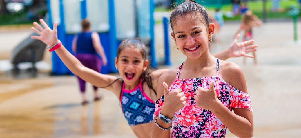 Two young girls in brightly coloured bathing suits, one posing with two thumbs up and the other behind her with her arms up in excitement in City Park Waterpark.
