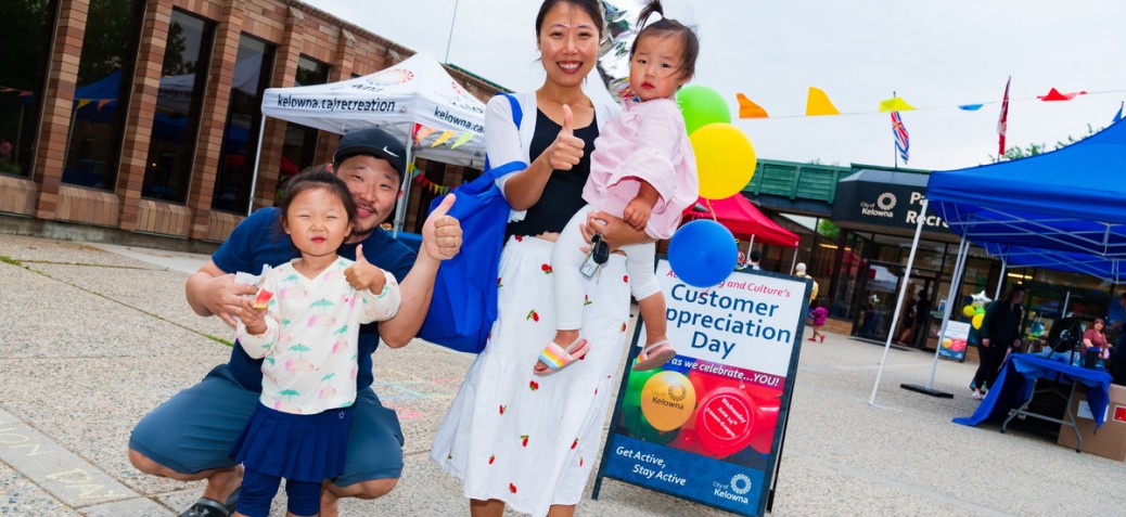 A young, smiling family stands in front of a Customer Appreciation Day sandwich board in front of PRC. The woman is holding a small child. The man is crouched beside another young child beside her. They are all giving a thumbs up to the camera.