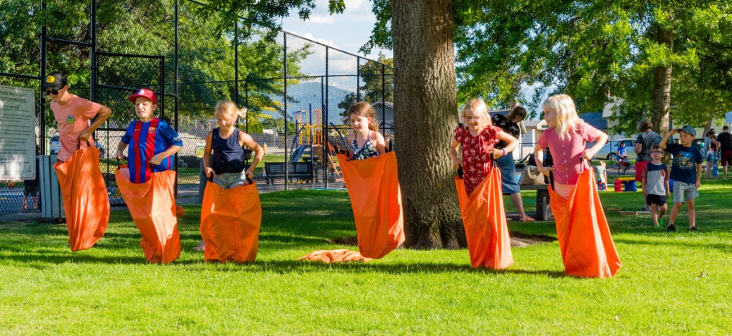 A group of six kids in orange sacks participating in a sack race on a sunny day at Hartwick Park.