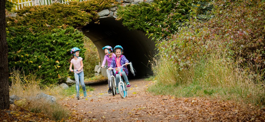 Three young girls ride bikes and scooters on a pathway in fall