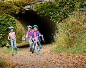 Three young girls ride bikes and scooters on an outdoor path