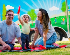 A smiling family looking at their young child holding up a giant checker piece on a sunny summer day